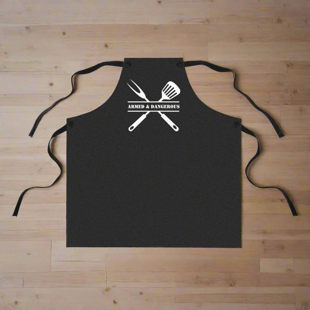 Black apron that says "Armed & Dangerous with a grill fork and spatula behind the text on a wooden surface
