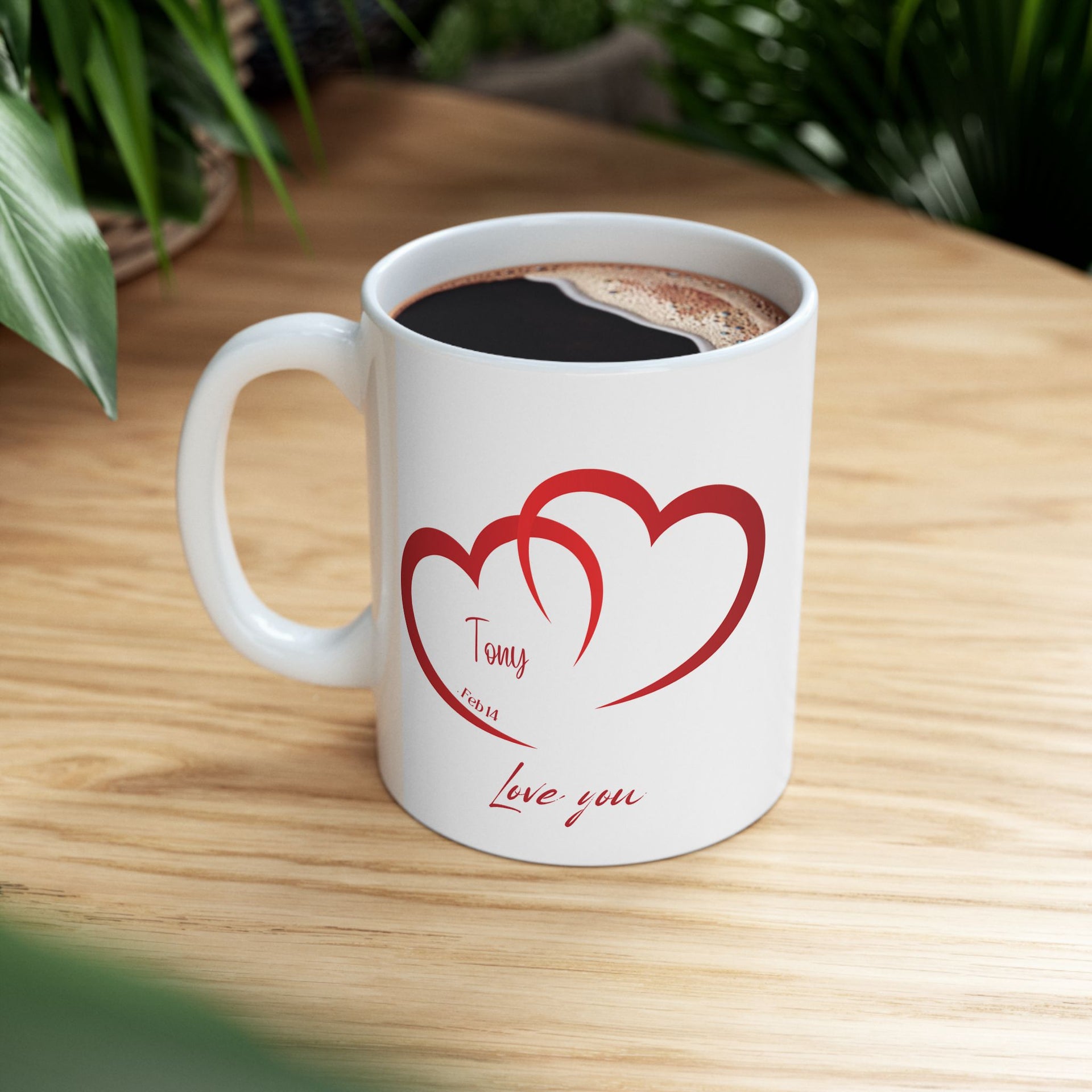 A white ceramic mug with two red heart shapes and the words 'Love you' printed on one side, placed on a wooden surface with a plant in the background.