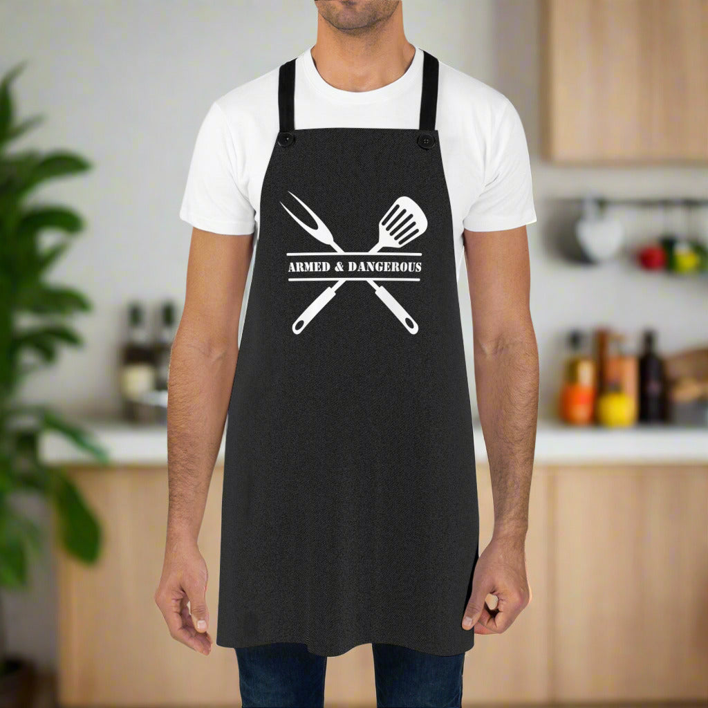 Man wearing a black apron that says "Armed and Dangerous". Behind the text is a grill fork and spatula
