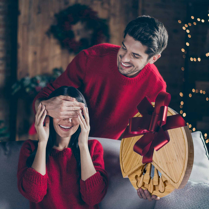 Man in red sweater holding a heart-shaped cutting board with a woman covering her eyes, surrounded by festive decorations.