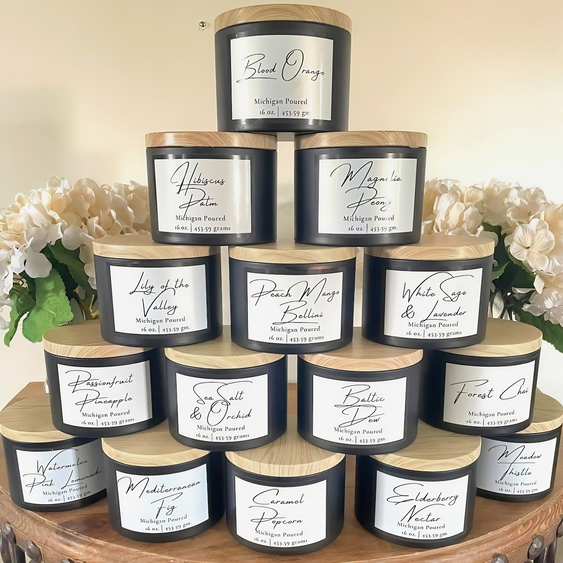 A collection of scented candles in various flavors, with black labels and wooden lids, displayed on a round wooden table with a white floral arrangement in the background.