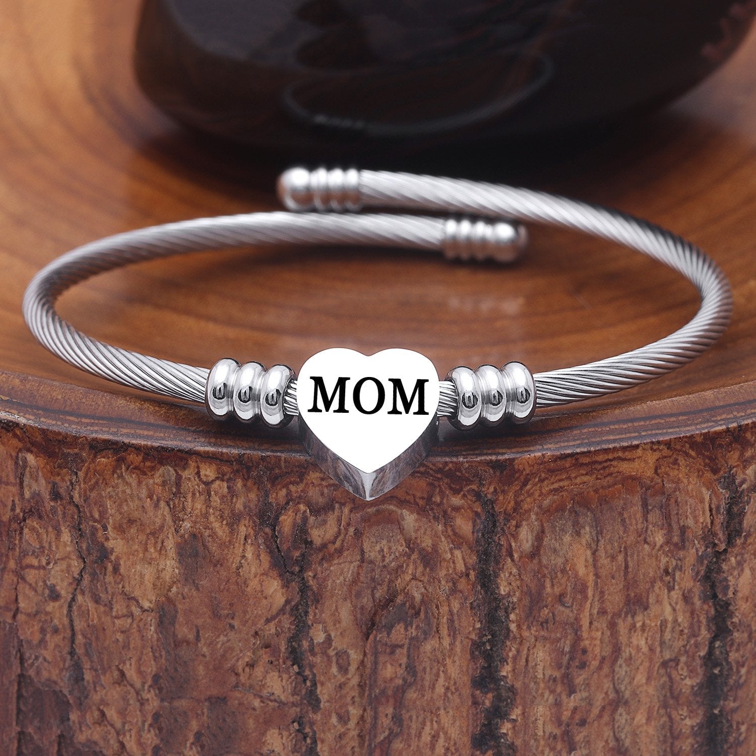 A stainless steel heart charm bracelet with the word 'MOM' inscribed on the heart, displayed on a wooden surface.