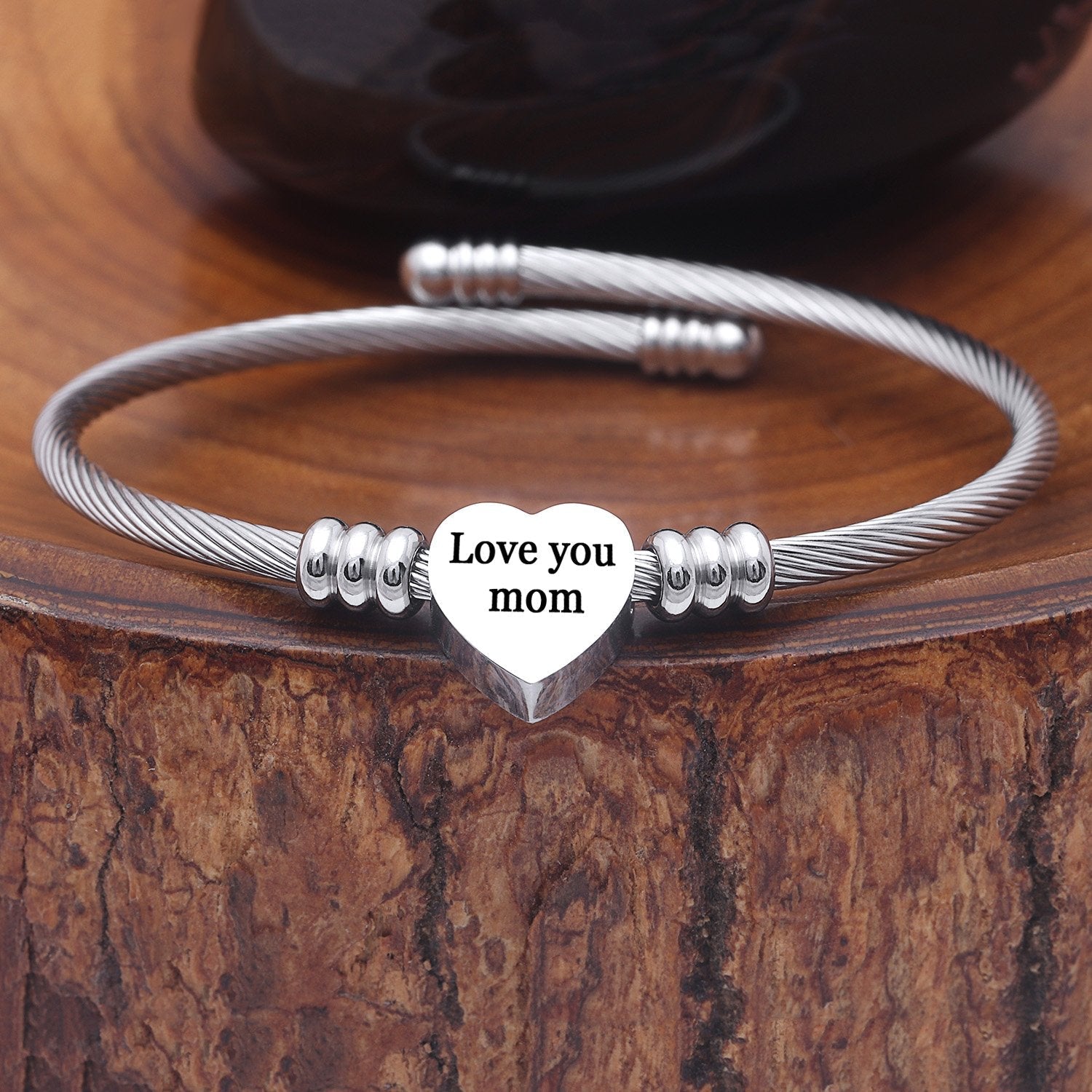 A stainless steel heart charm bracelet with the words 'Love you MOM' inscribed on the heart, displayed on a wooden surface.