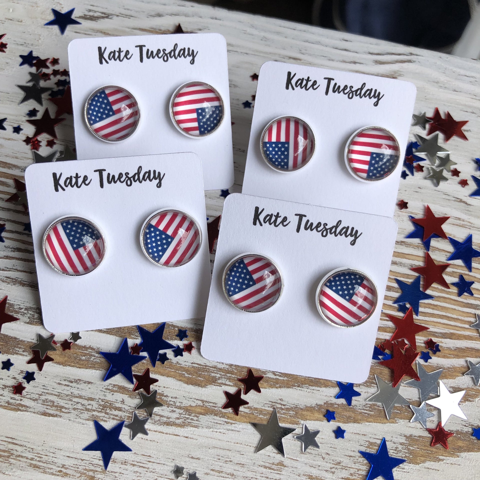 Four pairs of American flag earrings on 'Kate Tuesday' branded cards with decorative stars in the background.