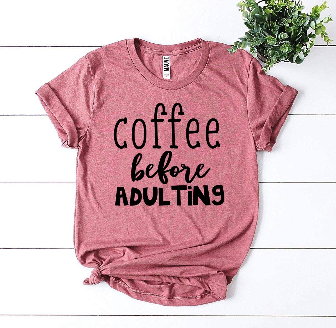 Pink t-shirt with 'coffee before adulting' text on a white wooden surface with a plant.