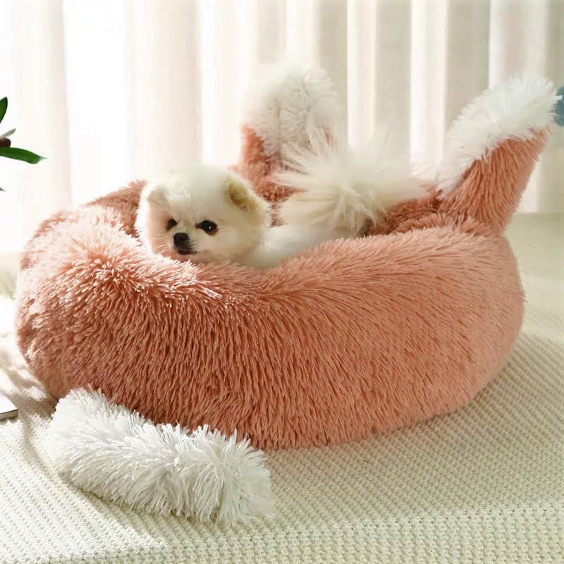 A small white dog resting on a plush pink pet bed with ears and tail design.