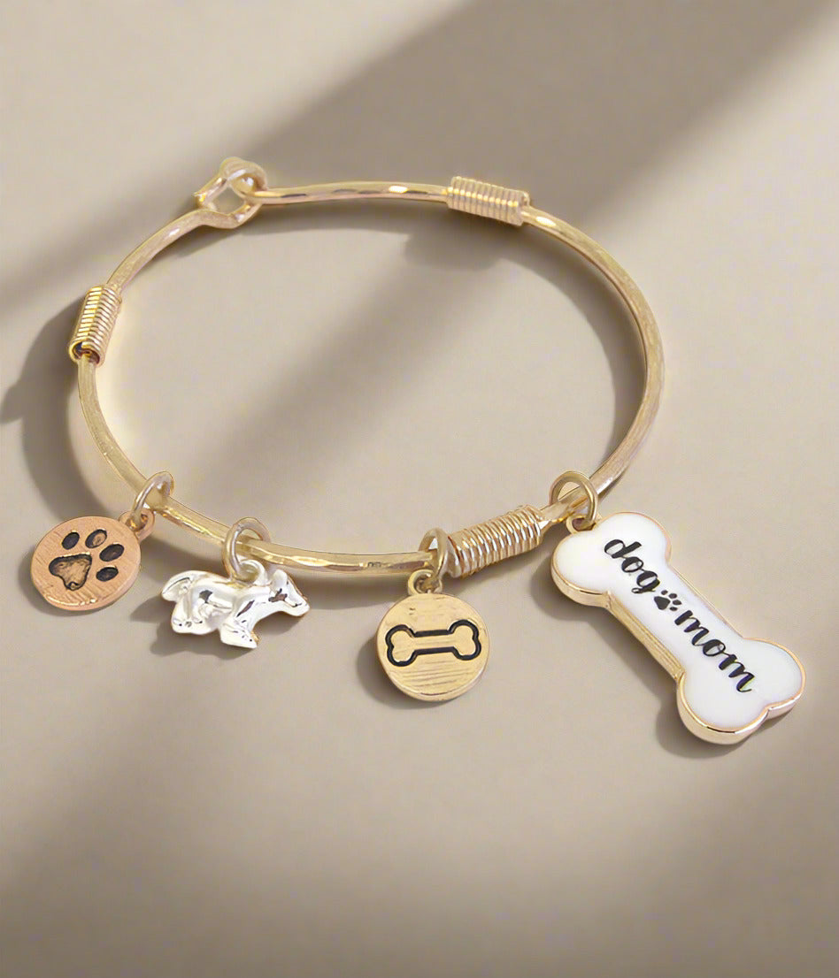 A gold-colored wire bangle bracelet with multiple charms including a paw print, a bone, and a tag with the text 'dog mom'.