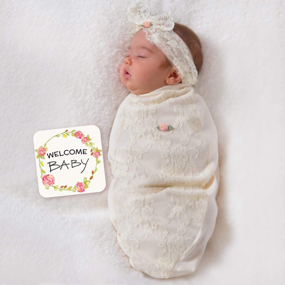 A newborn baby swaddled in a lace-patterned blanket with a matching headband, lying on a white surface, with a 'WELCOME BABY' announcement card beside it.