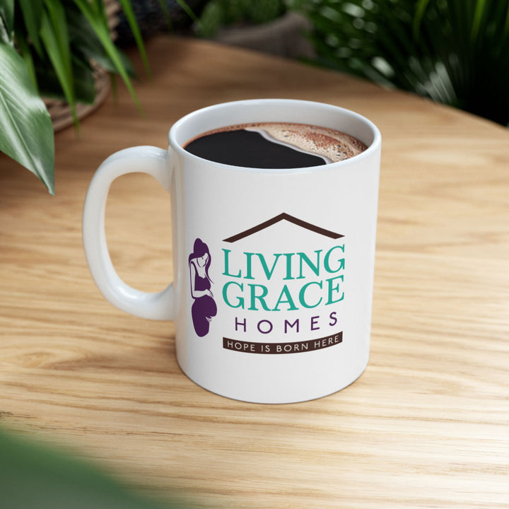 White ceramic mug filled with coffee, featuring the Living Grace Homes logo with a purple silhouette of a mother and baby and the phrase “Hope is born here,” displayed on a wooden table surrounded by green plants. Inspirational nonprofit coffee mug supporting mothers in need.