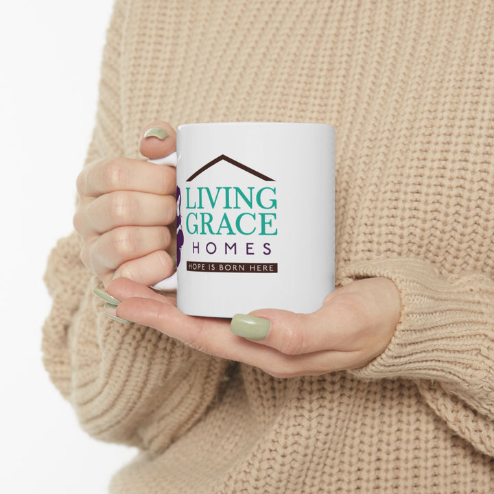 Woman in a cozy beige sweater holding a white Living Grace Homes mug featuring the logo and the phrase “Hope is born here.” Inspirational nonprofit coffee mug supporting mothers and families in need.