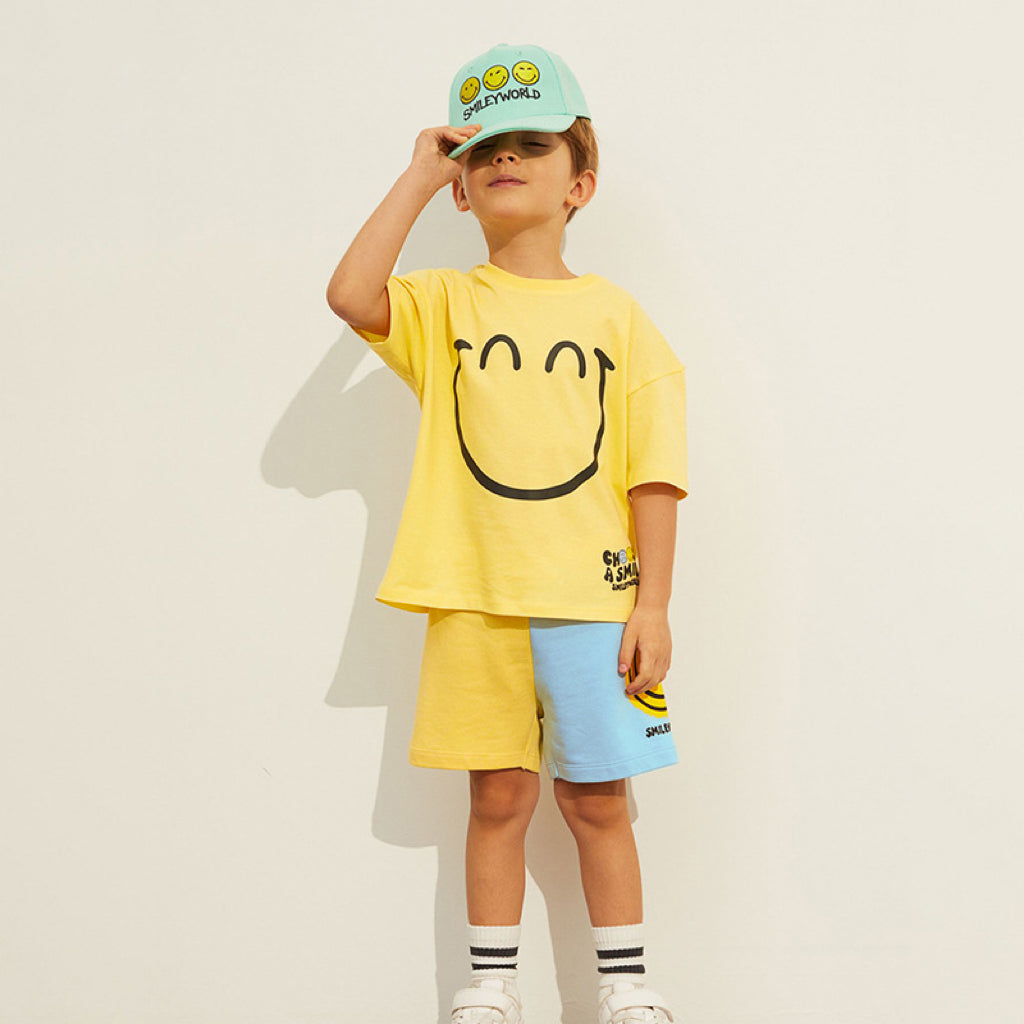 Child wearing a yellow smiley face t-shirt, blue shorts, and a matching cap on a plain background
