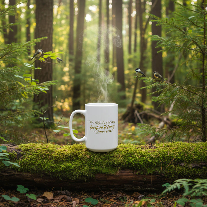 White mug with gold text 'You didn't choose birdwatching, it chose you.' on a forest log, with birds watching
