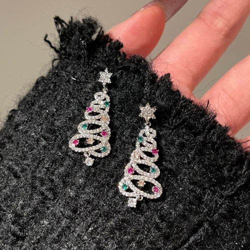 Silver Christmas tree earrings with gemstones held against a textured black background.