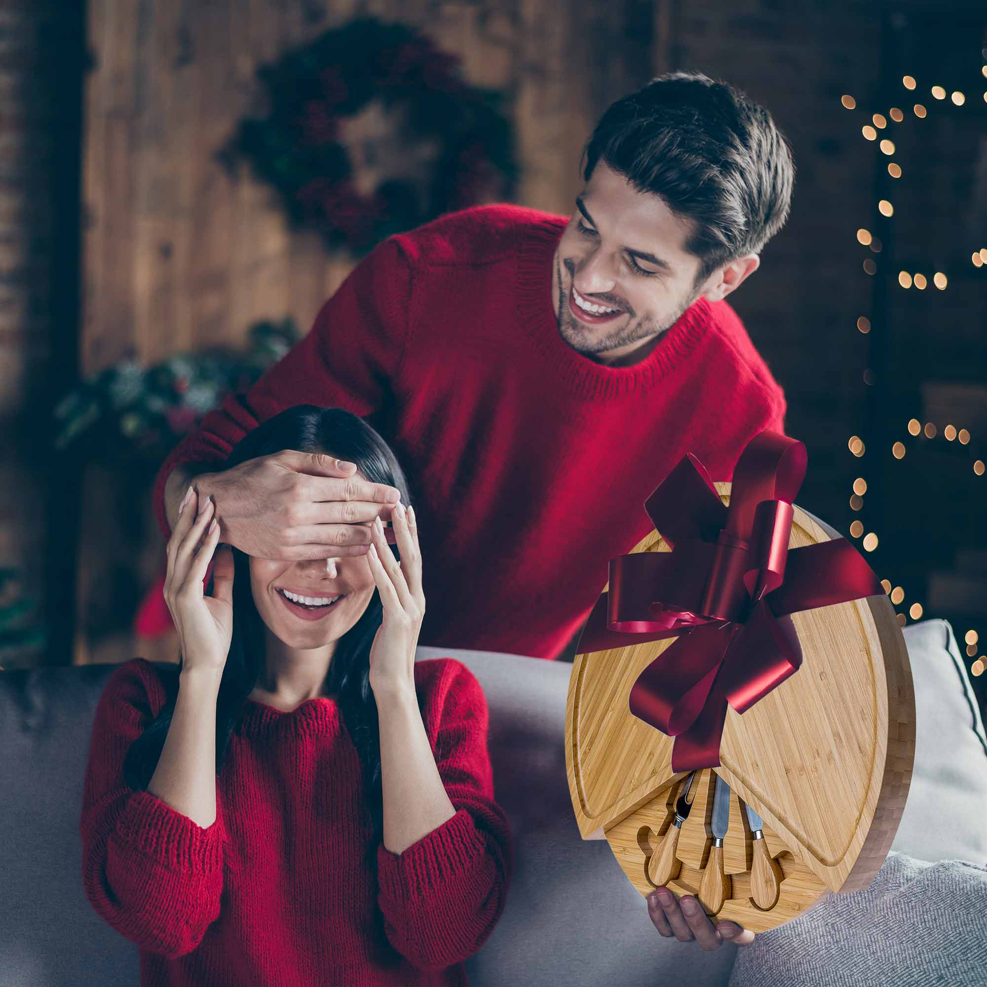 Man in red sweater holding a heart-shaped cutting board with a woman covering her eyes, surrounded by festive decorations.