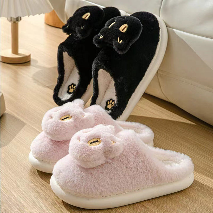 Pair of black cat-shaped slippers on a wooden floor with a white couch in the background.