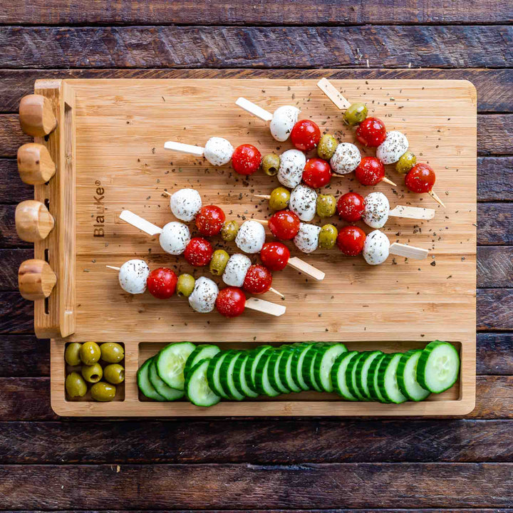 Wooden cutting board with skewers of olives, tomatoes, and mozzarella balls, accompanied by sliced cucumbers.