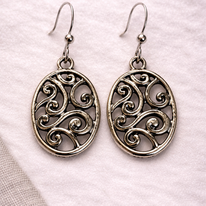 Silver oval filigree dangle earrings with intricate swirling metal design on soft neutral background
