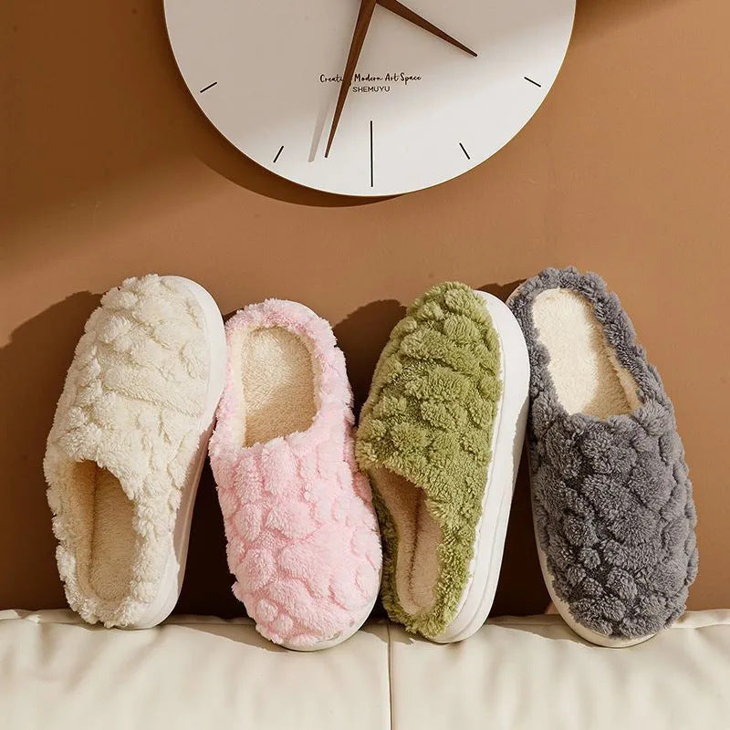 Four pairs of textured slippers in white, pink, green, and gray on a beige surface with a clock in the background.