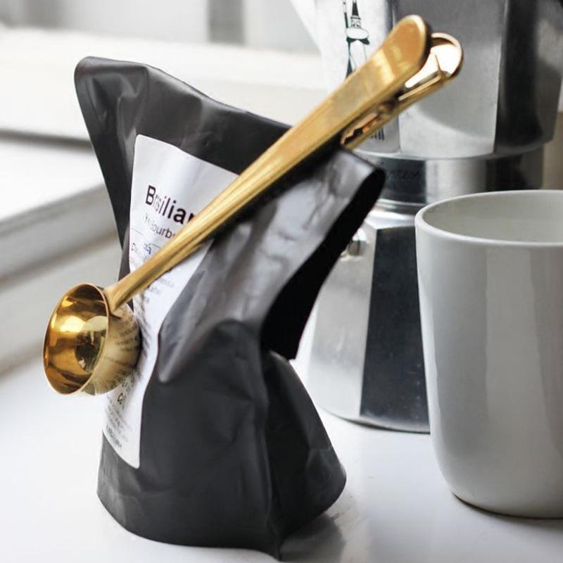 Gold stainless steel coffee scoop clipped onto coffee bag beside cup