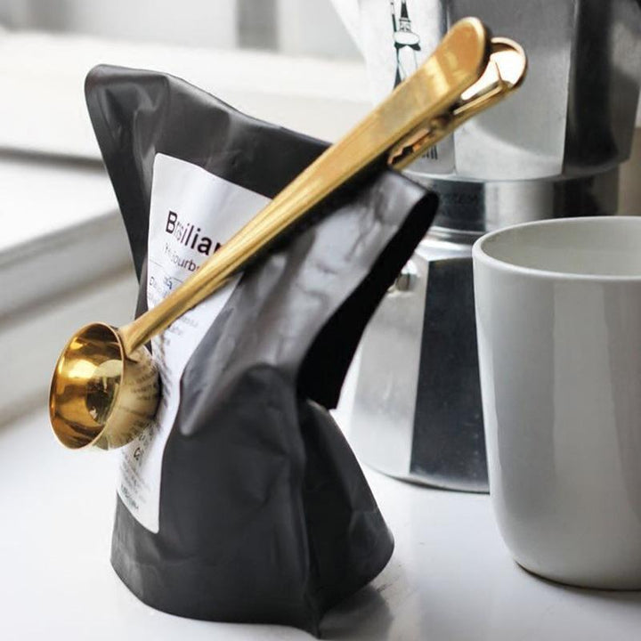 Gold stainless steel coffee scoop clipped onto coffee bag beside cup