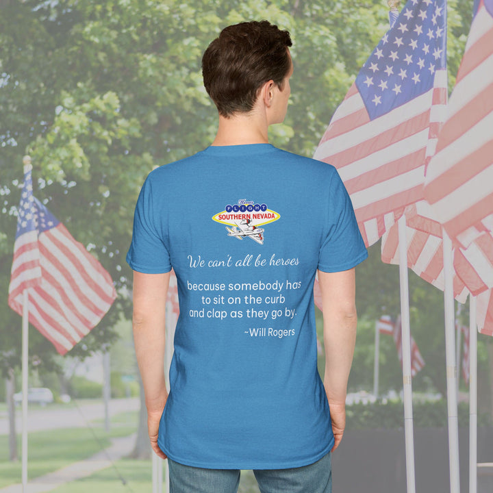 Person wearing a blue t-shirt with text and an airplane graphic, standing in front of American flags.