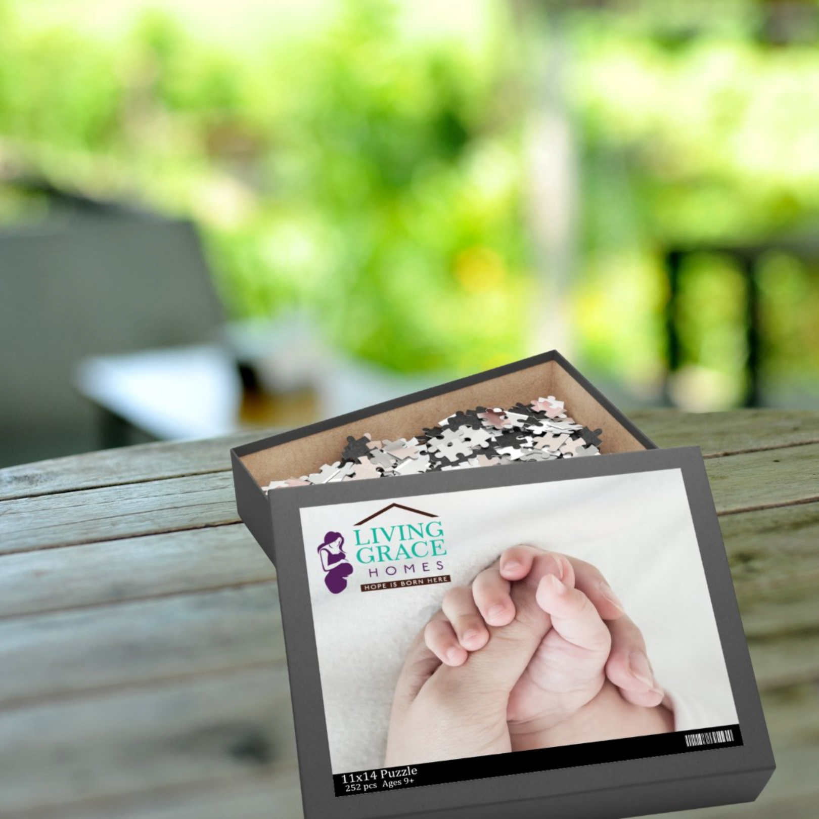 An open jigsaw puzzle box with pieces displayed, showing the image of two hands holding each other. The box is labeled with the brand 'LIVING GRACE'.
