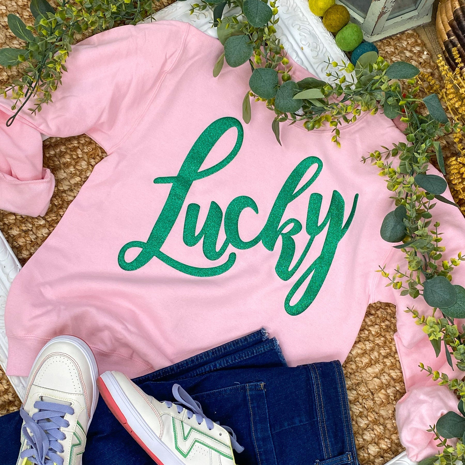 Pink sweatshirt with 'Lucky' text, white sneakers, and denim jeans on a decorative surface with greenery and colorful pom-poms.