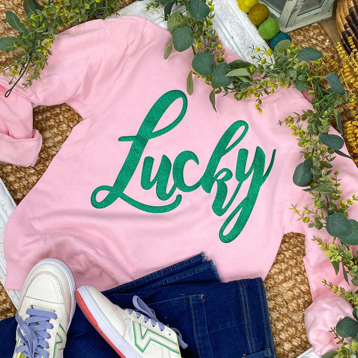 Pink sweatshirt with 'Lucky' text, white sneakers, and denim jeans on a decorative surface with greenery and colorful pom-poms.