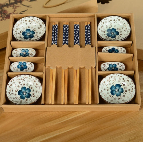 Set of ceramic bowls with floral designs and chopsticks in a cardboard box on a wooden surface.