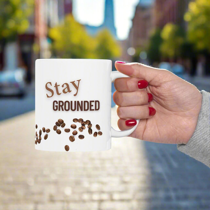 woman holding a while mug that says "stay grounded". Mug also has coffee beans scattered around as a design element.