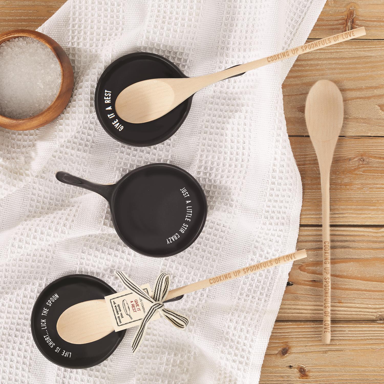 Set of wooden cooking spoons with engraved handles reading “Cooking Up Spoonfuls of Love,” paired with black ceramic spoon rests featuring playful quotes like “Give It a Rest,” “Just a Little Stir Crazy,” and “Life is Short, Lick the Spoon,” displayed on a rustic kitchen towel, unique cooking gift set for home chefs.