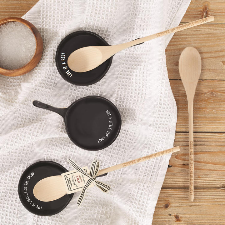 Set of wooden cooking spoons with engraved handles reading “Cooking Up Spoonfuls of Love,” paired with black ceramic spoon rests featuring playful quotes like “Give It a Rest,” “Just a Little Stir Crazy,” and “Life is Short, Lick the Spoon,” displayed on a rustic kitchen towel, unique cooking gift set for home chefs.