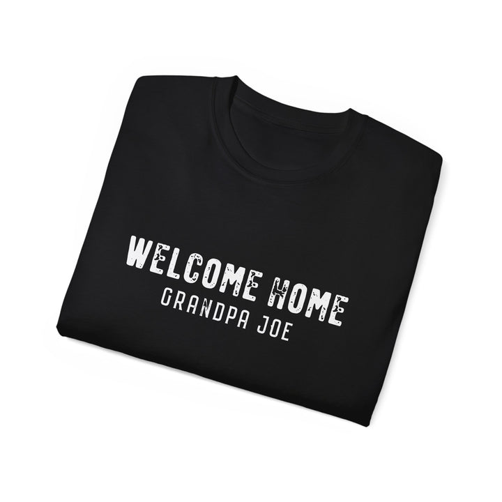 A folded black t-shirt with 'WELCOME HOME GRANDPA JOE' printed in distressed white text on the front, displayed on a clean white background.