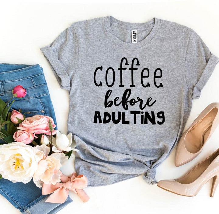 Gray t-shirt with 'coffee before adulting' text, paired with jeans, shoes, and flowers on a white background