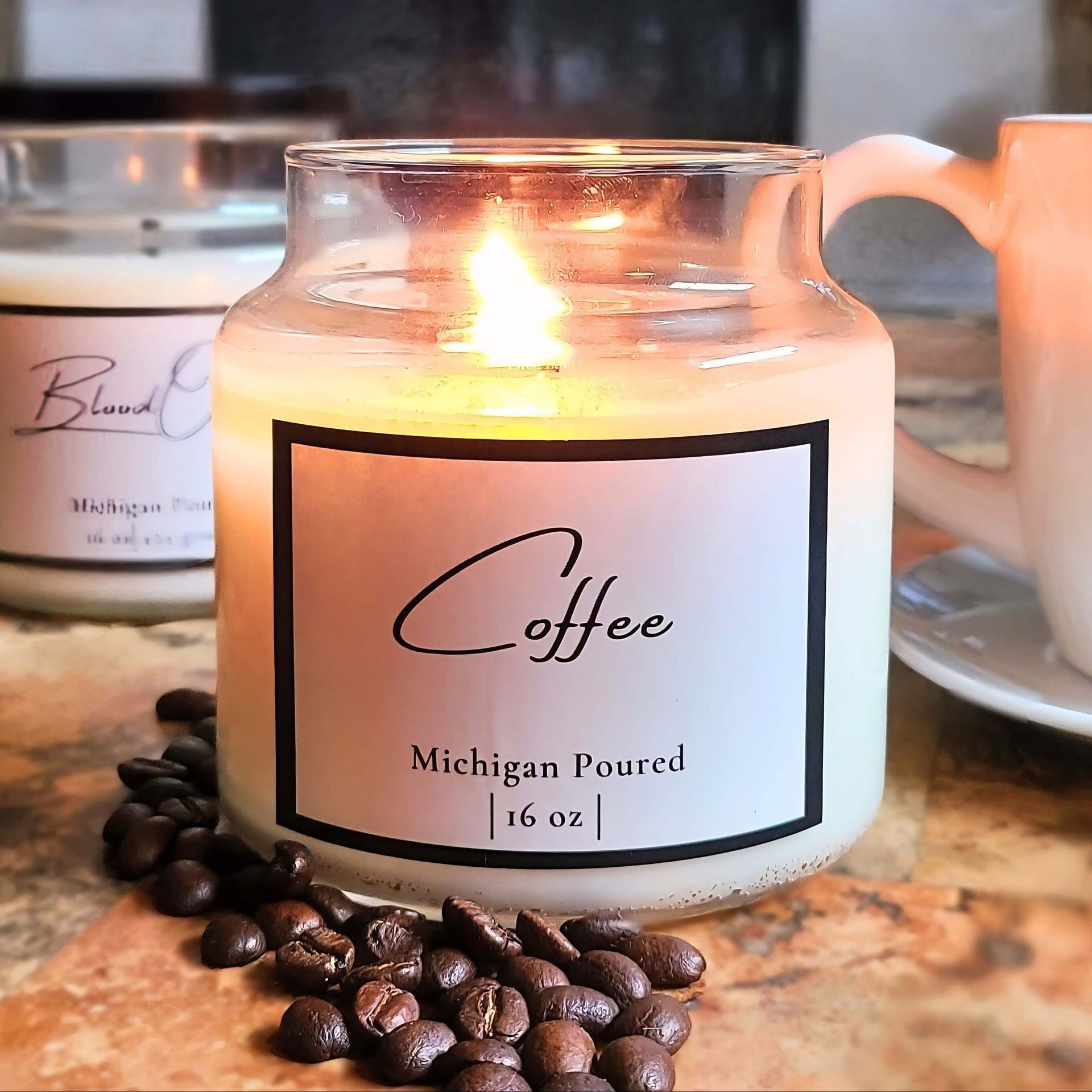 Candle labeled 'Coffee' with coffee beans on a surface, with another candle and mug in the background.