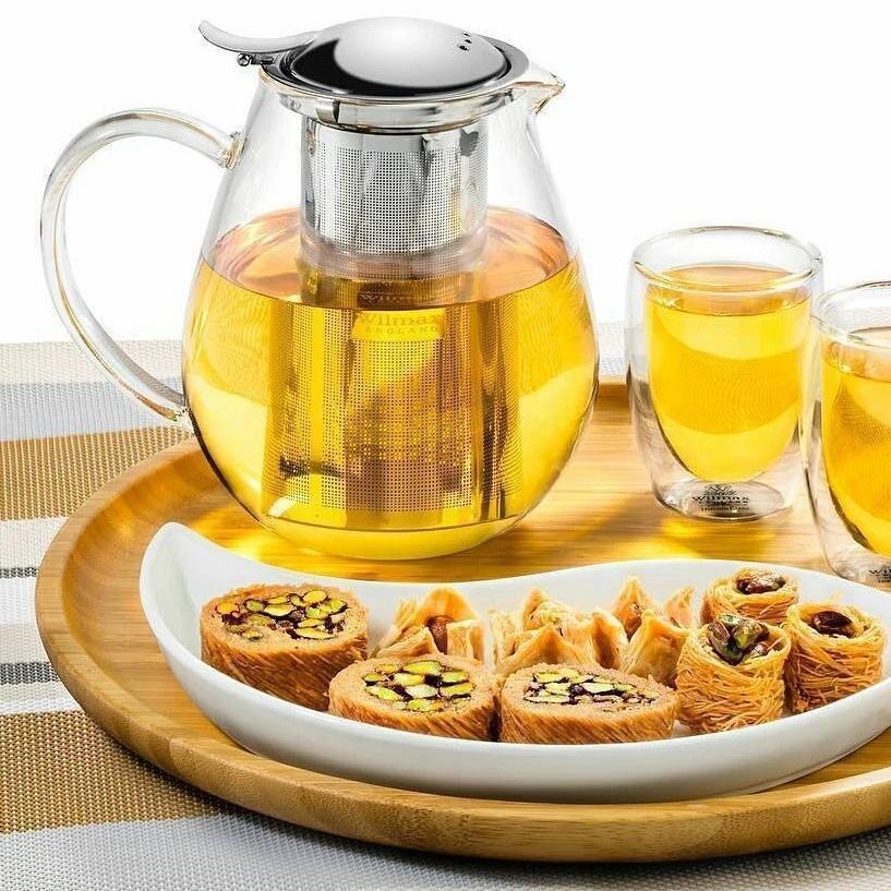Glass teapot with stainless steel infuser filled with tea, served with clear glass cups and pastries on a wooden tray