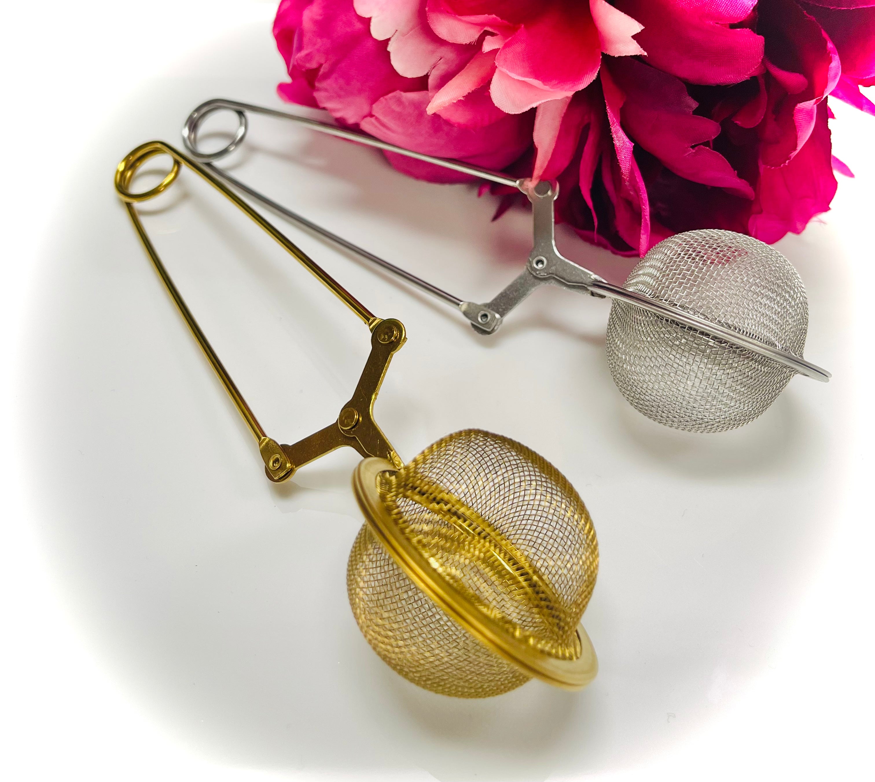 Gold and silver stainless steel tea infuser ball tongs with fine mesh design beside pink flowers