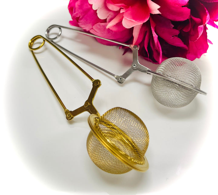 Gold and silver stainless steel tea infuser ball tongs with fine mesh design beside pink flowers
