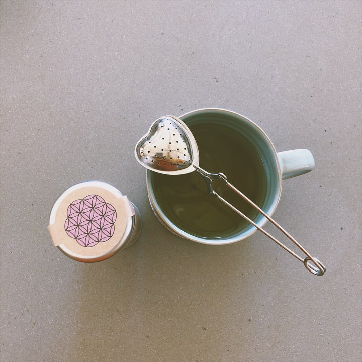 Heart-shaped stainless steel tea infuser resting on a cup of tea beside a jar of loose leaf tea