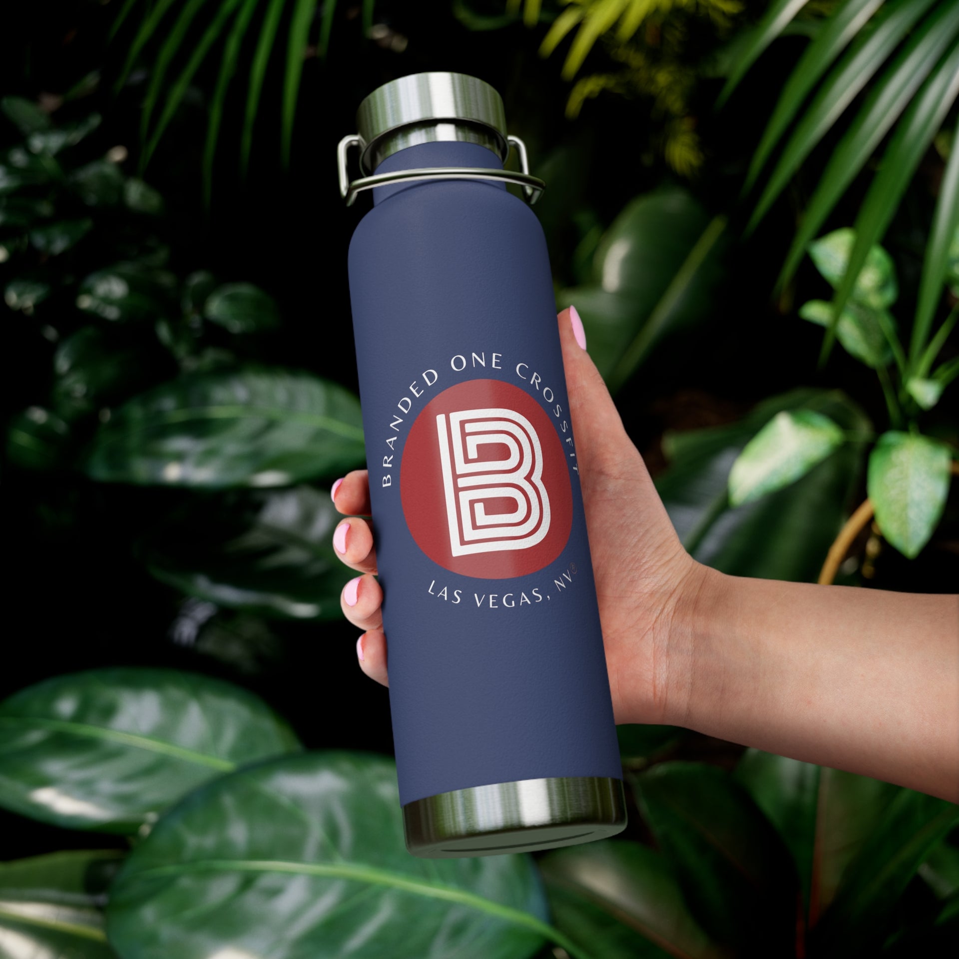 A navy blue copper vacuum insulated bottle with a white base and red Branded One Crossfit logo, being held by a hand against a backdrop of green foliage.