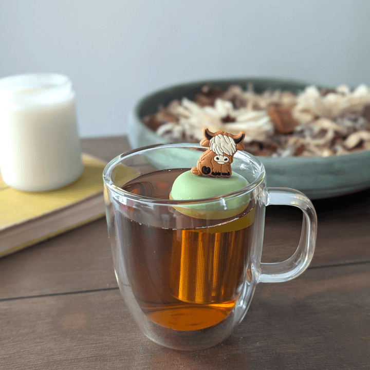 Floating Adult Highland Cow Tea Infuser in a clear mug of tea with a green lid, perfect for loose leaf tea.