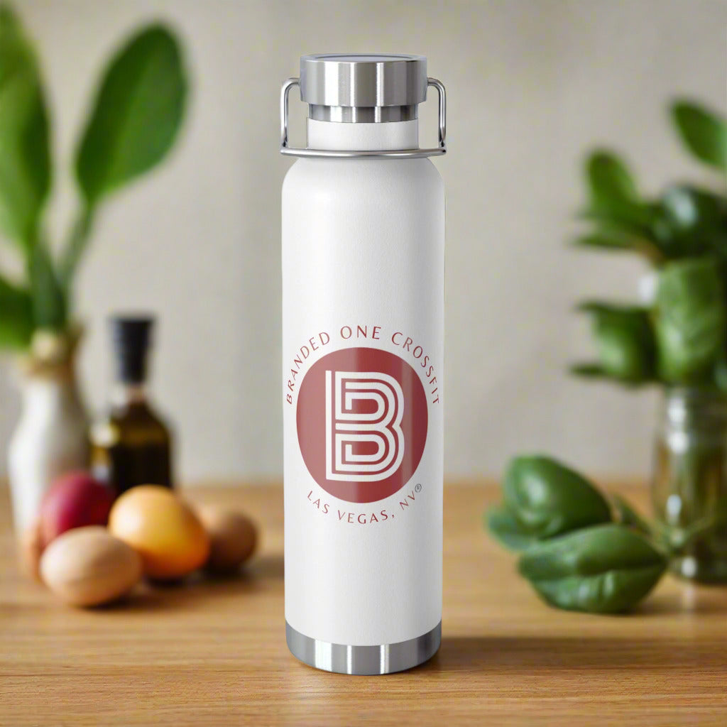 A white copper vacuum insulated bottle with a white base and red Branded One Crossfit logo, being held by a hand against a backdrop of green foliage.